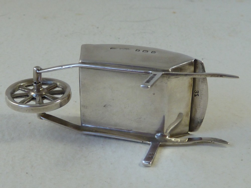 A miniature sterling silver stamp case shaped like a garden wheelbarrow with a clear glass window displaying a pink postage stamp. View VI