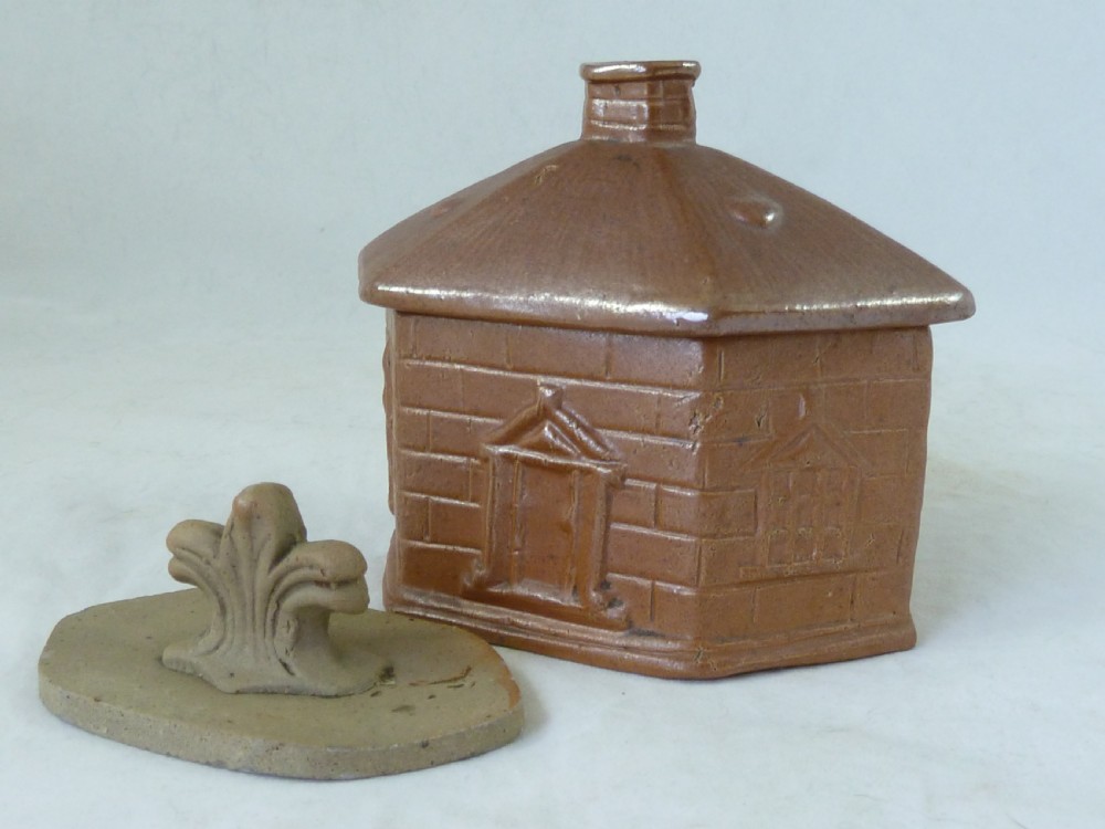 A warm brown salt-glazed stoneware tobacco jar shaped like a toll house cottage alongside its separate internal clay presser.