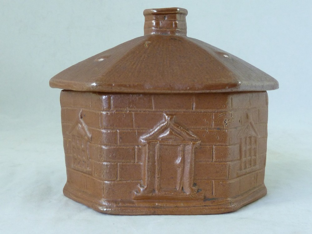 A warm brown salt-glazed stoneware tobacco jar shaped like a toll house cottage alongside its separate internal clay presser. View II