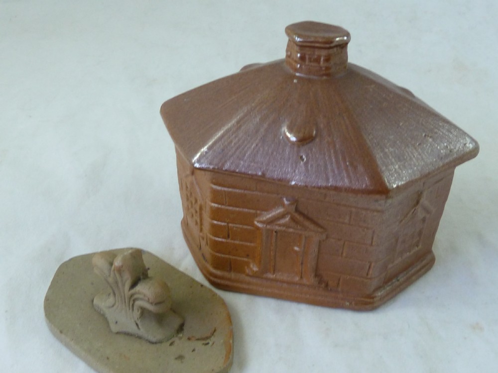 A warm brown salt-glazed stoneware tobacco jar shaped like a toll house cottage alongside its separate internal clay presser. View III