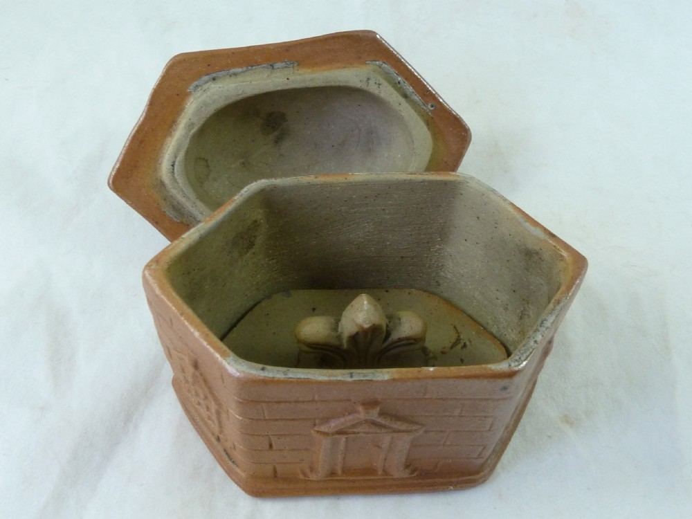 A warm brown salt-glazed stoneware tobacco jar shaped like a toll house cottage alongside its separate internal clay presser. View V