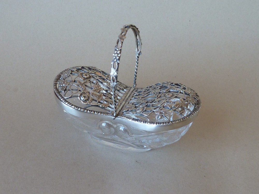 An oval clear cut glass bowl with a pierced silver floral lid and an overhead swing handle.