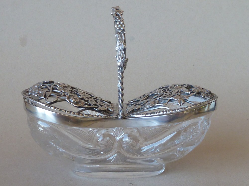 An oval clear cut glass bowl with a pierced silver floral lid and an overhead swing handle. View II