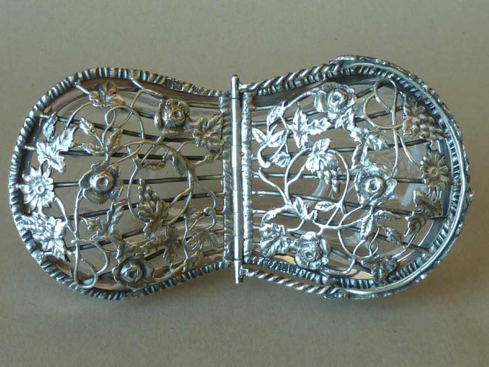 An oval clear cut glass bowl with a pierced silver floral lid and an overhead swing handle. View V