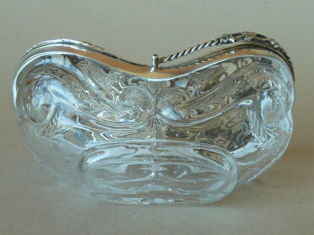An oval clear cut glass bowl with a pierced silver floral lid and an overhead swing handle. View VI