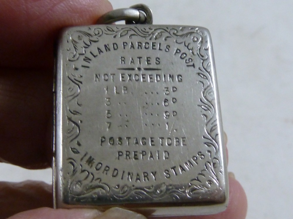 A rectangular Victorian silver-coloured metal stamp case fob featuring engraved scrollwork borders and etched text displaying inland parcel post rates.