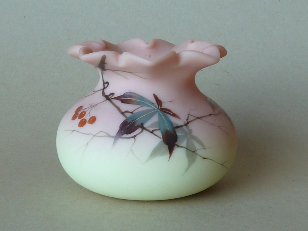A small Victorian Burmese satin glass vase with a crimped rim, shading from yellow at the base to pink at the top, decorated with hand-painted leaves and berries.