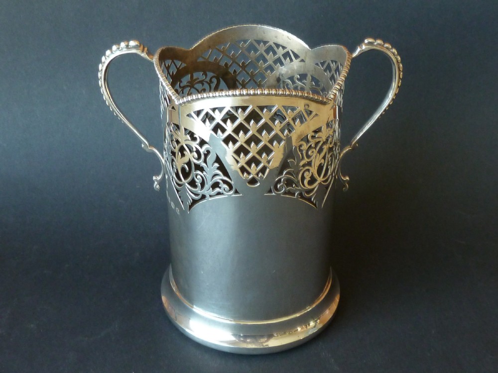 A solid silver magnum wine bottle holder featuring pierced fretwork detail, two beaded scroll handles, and British hallmarks on the side.