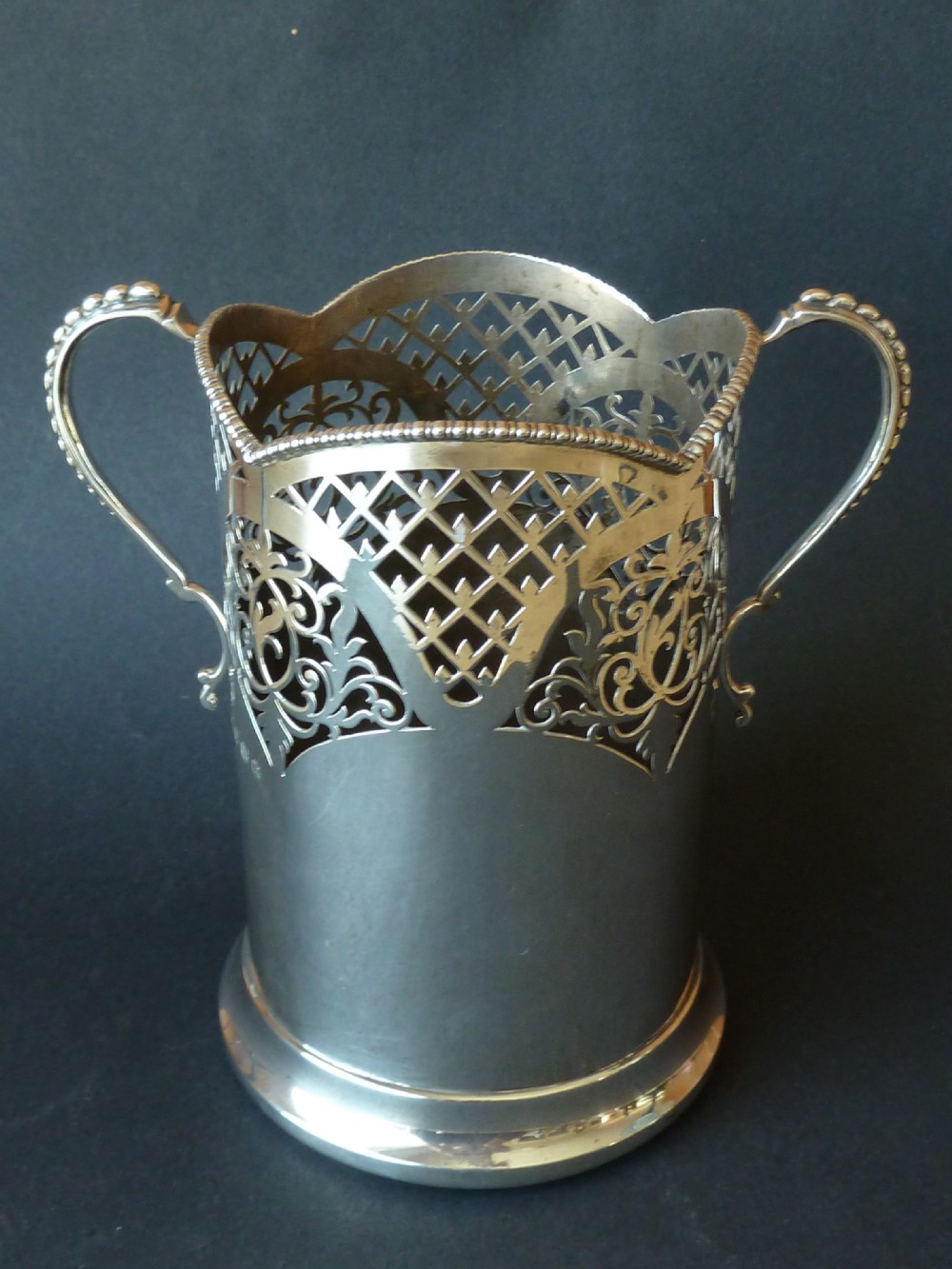 A solid silver magnum wine bottle holder featuring pierced fretwork detail, two beaded scroll handles, and British hallmarks on the side. View II