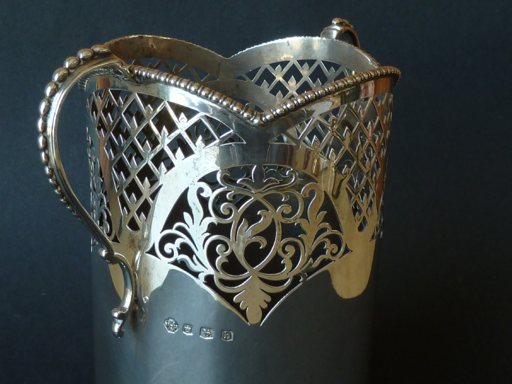 A solid silver magnum wine bottle holder featuring pierced fretwork detail, two beaded scroll handles, and British hallmarks on the side. View III