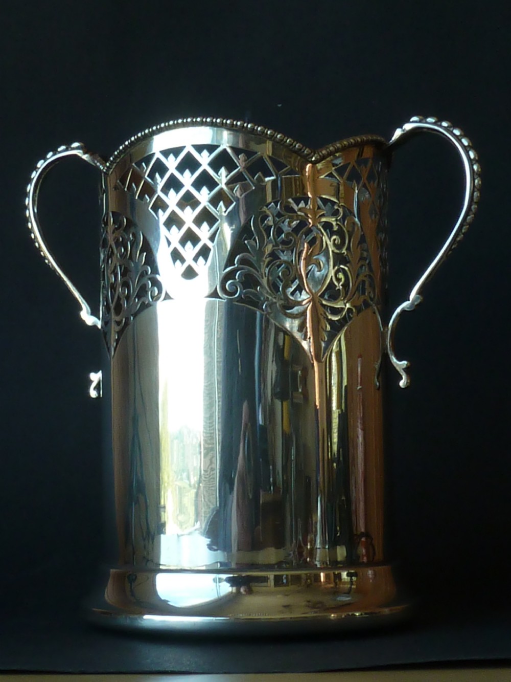 A solid silver magnum wine bottle holder featuring pierced fretwork detail, two beaded scroll handles, and British hallmarks on the side. View VI