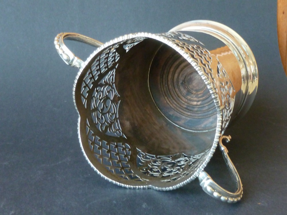 A solid silver magnum wine bottle holder featuring pierced fretwork detail, two beaded scroll handles, and British hallmarks on the side. View VII