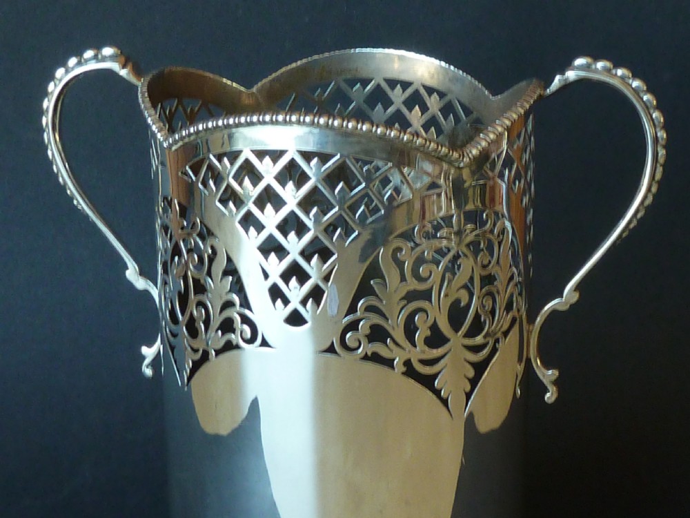 A solid silver magnum wine bottle holder featuring pierced fretwork detail, two beaded scroll handles, and British hallmarks on the side. View VIII
