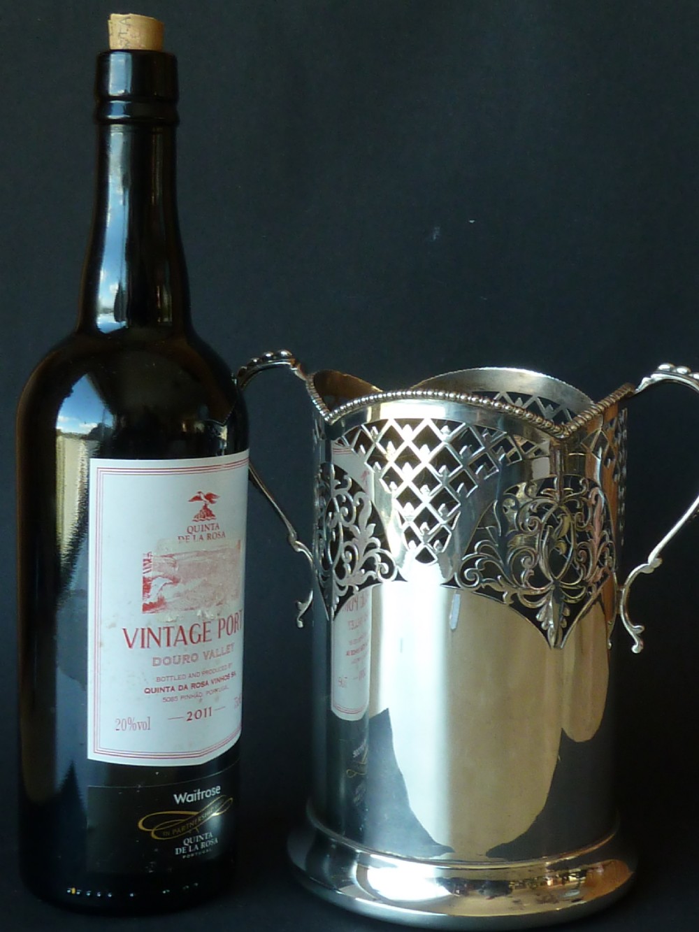 A solid silver magnum wine bottle holder featuring pierced fretwork detail, two beaded scroll handles, and British hallmarks on the side. View IX