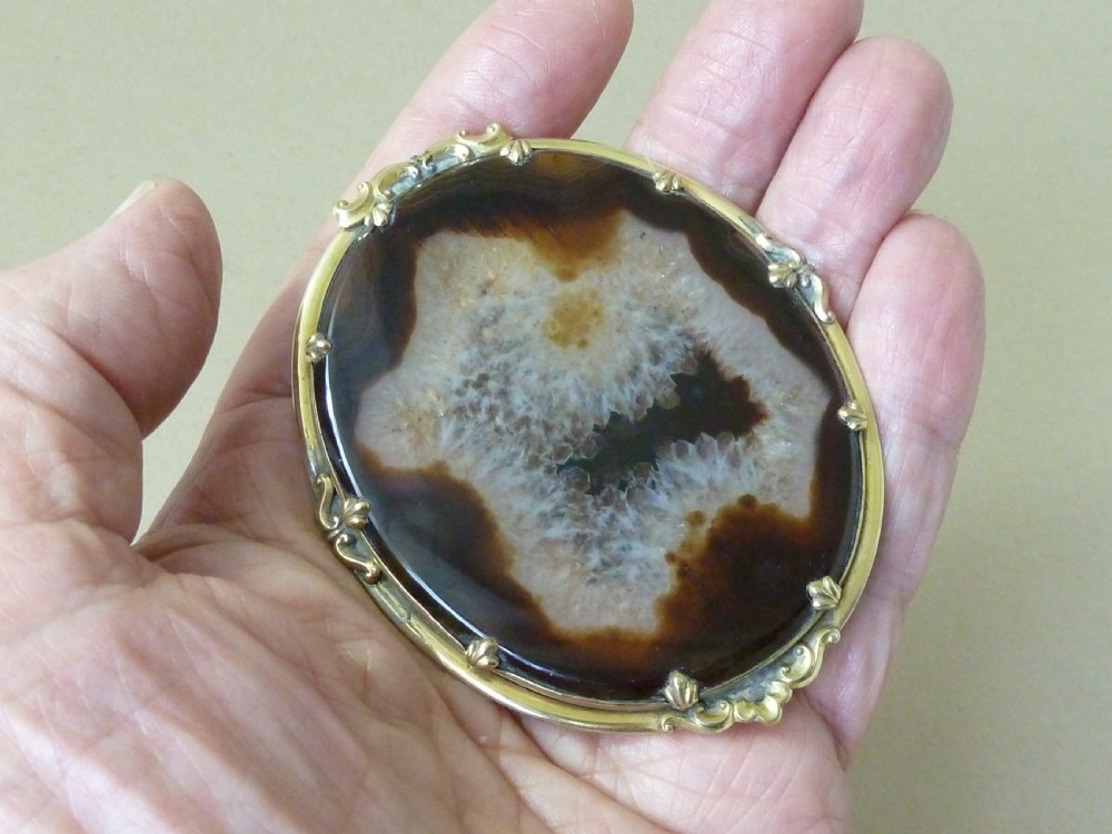 An oval Victorian brooch featuring a translucent brown and white patterned agate stone set within a gold-toned metal frame with decorative prongs. View VI