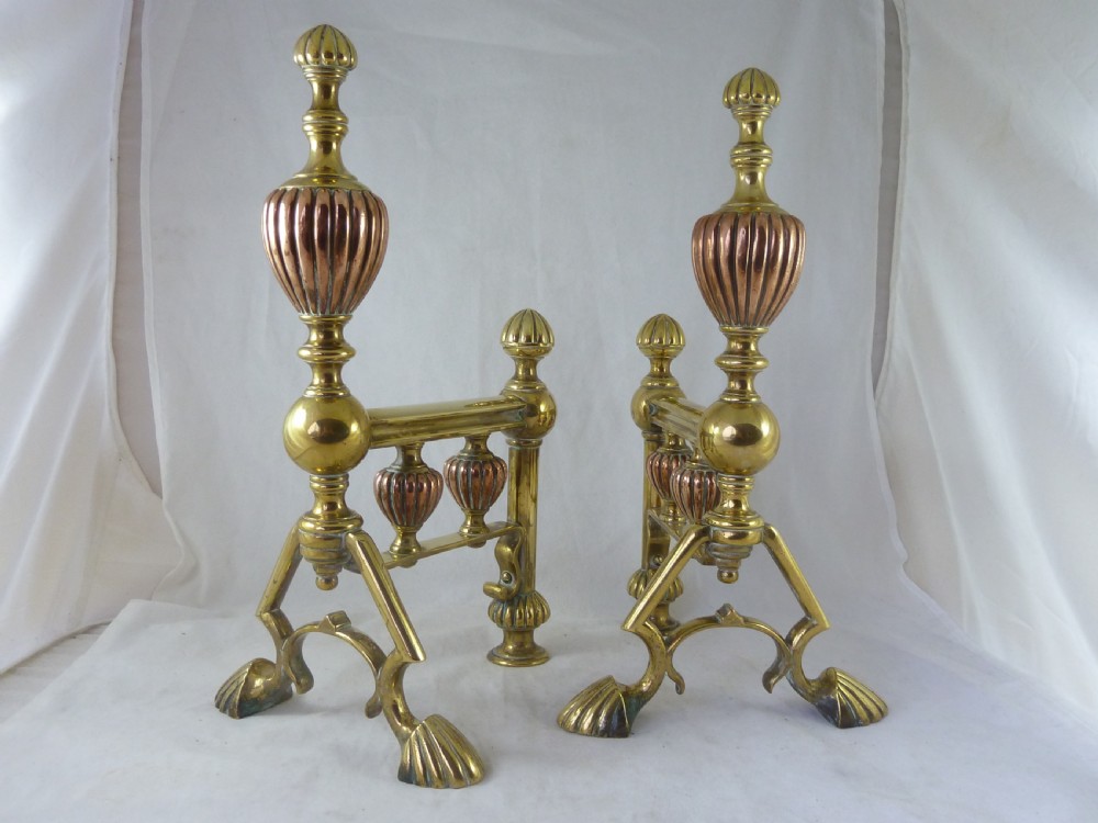 A pair of Victorian brass and copper fire dogs featuring ribbed urn-shaped finials, turned posts, and claw feet.