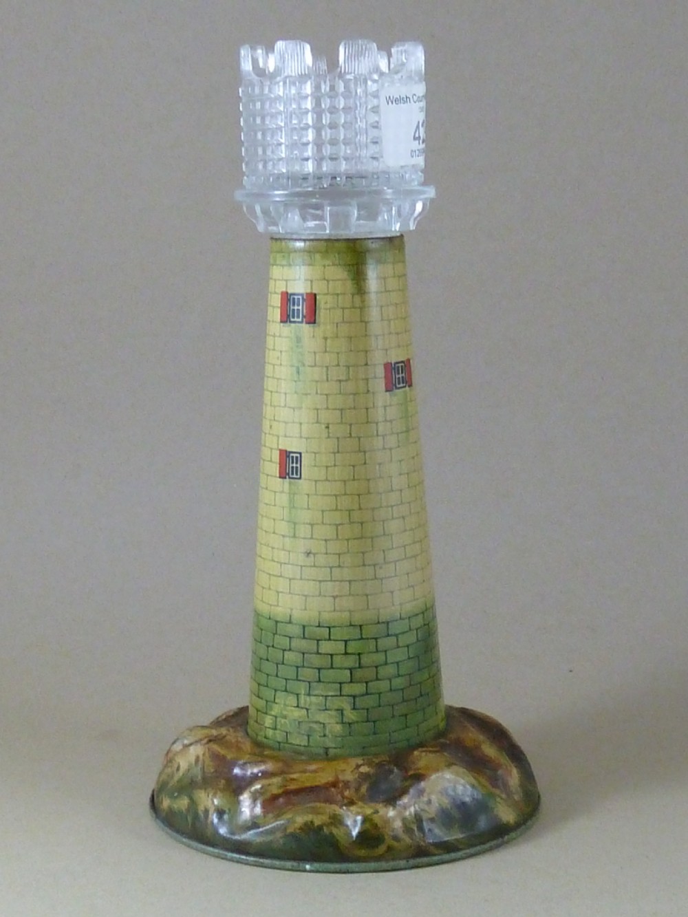 A Victorian lithographed tin candle holder shaped like a lighthouse with a clear pressed glass lantern shade. View V