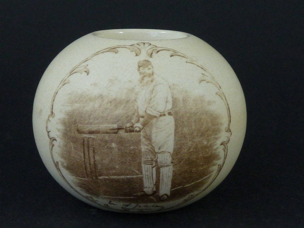A spherical cream ceramic match striker featuring a sepia transfer print of cricketer W.G. Grace holding a bat within a scrollwork frame.