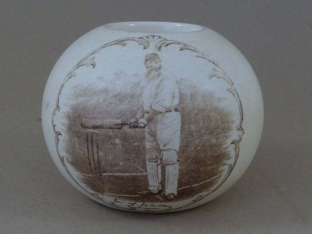 A spherical cream ceramic match striker featuring a sepia transfer print of cricketer W.G. Grace holding a bat within a scrollwork frame. View II