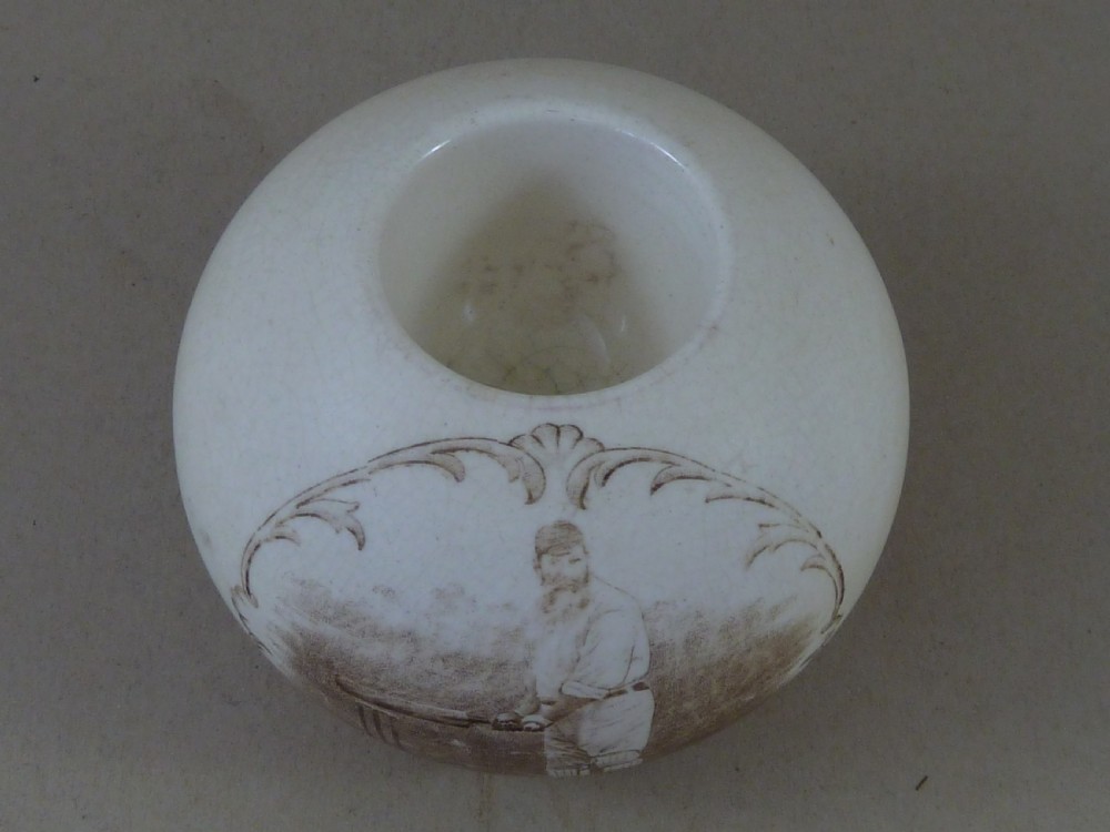 A spherical cream ceramic match striker featuring a sepia transfer print of cricketer W.G. Grace holding a bat within a scrollwork frame. View VI