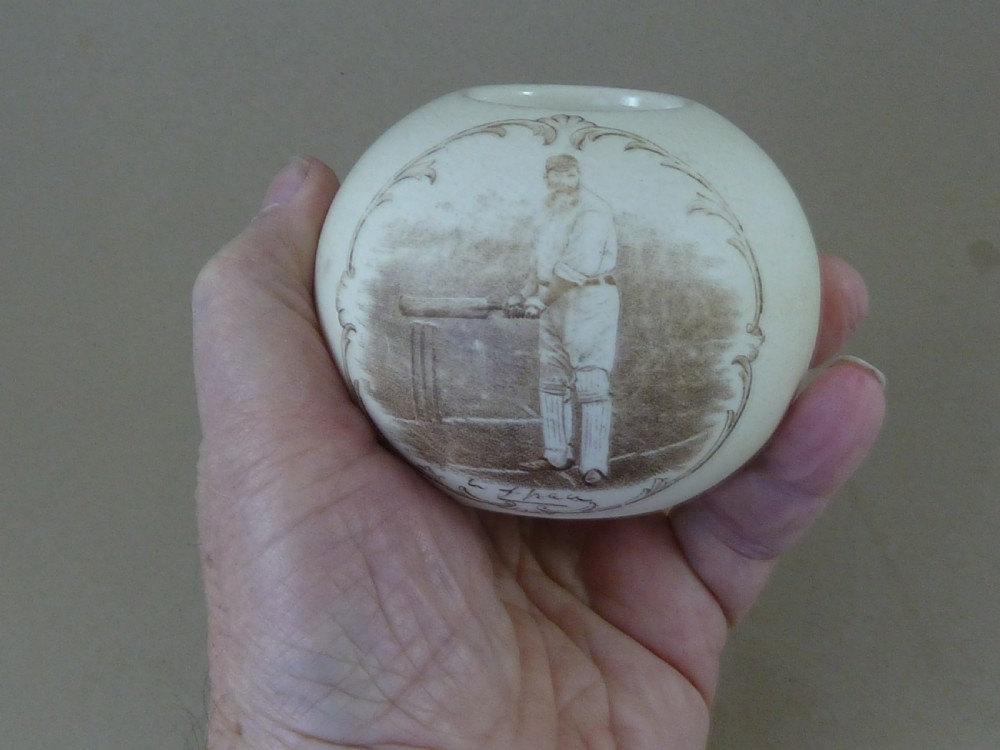 A spherical cream ceramic match striker featuring a sepia transfer print of cricketer W.G. Grace holding a bat within a scrollwork frame. View VII