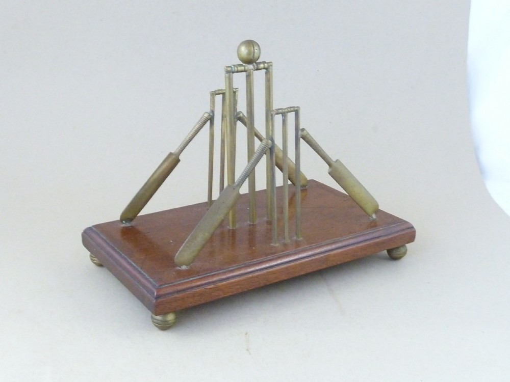 An antique brass letter rack shaped as miniature cricket bats, wickets, and a ball mounted on a rectangular mahogany wooden base with small bun feet.