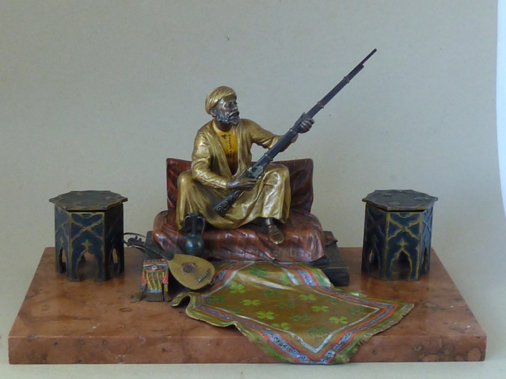 A cold-painted Vienna bronze inkstand featuring a seated figure holding a long firearm mounted on a reddish marble slab alongside twin octagonal inkwell boxes.