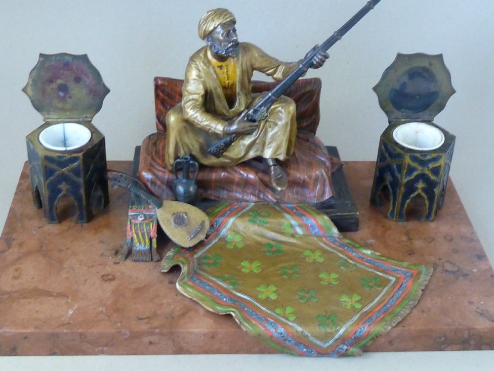 A cold-painted Vienna bronze inkstand featuring a seated figure holding a long firearm mounted on a reddish marble slab alongside twin octagonal inkwell boxes. View V