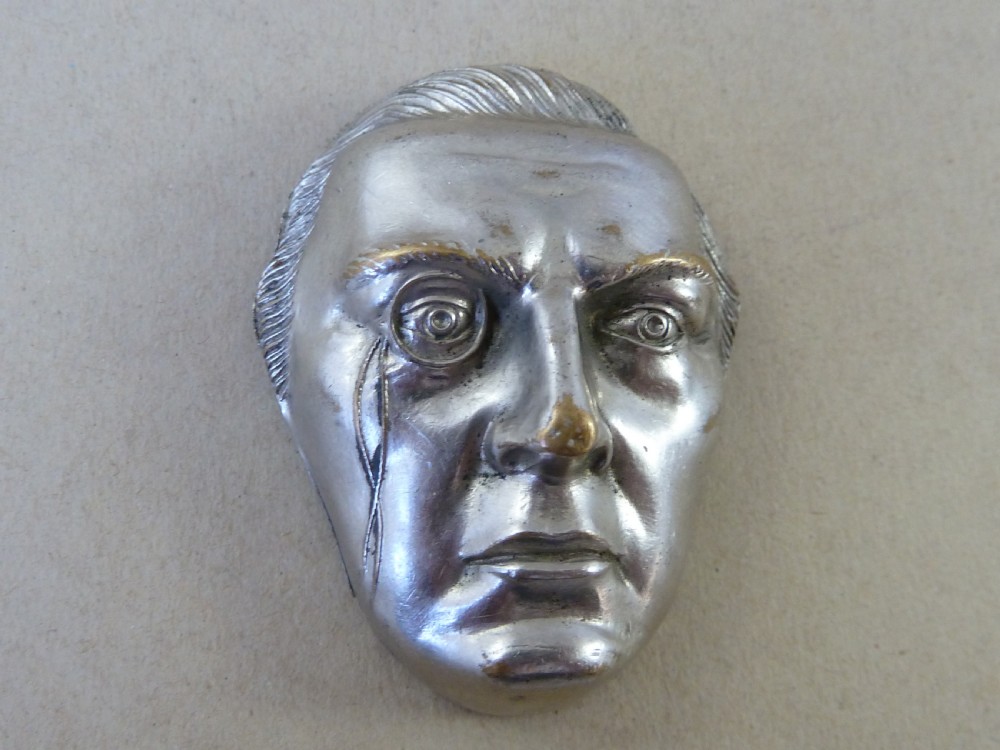 A nickel-plated metal vesta case cast in the high-relief shape of a mans face wearing a monocle.