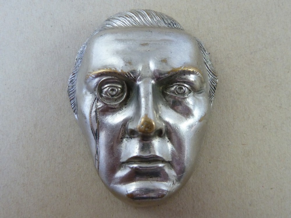 A nickel-plated metal vesta case cast in the high-relief shape of a mans face wearing a monocle. View II