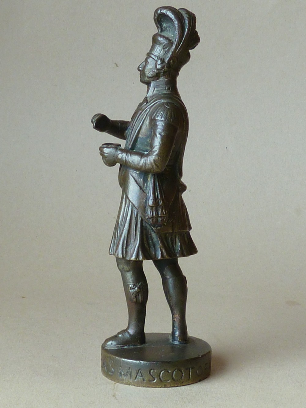 A dark bronze figure depicting a Scottish Highlander in military uniform standing on a circular base inscribed with text.