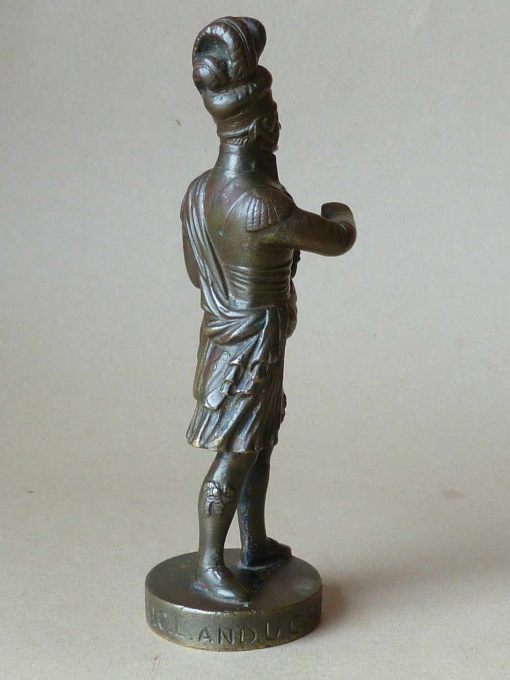 A dark bronze figure depicting a Scottish Highlander in military uniform standing on a circular base inscribed with text. View II