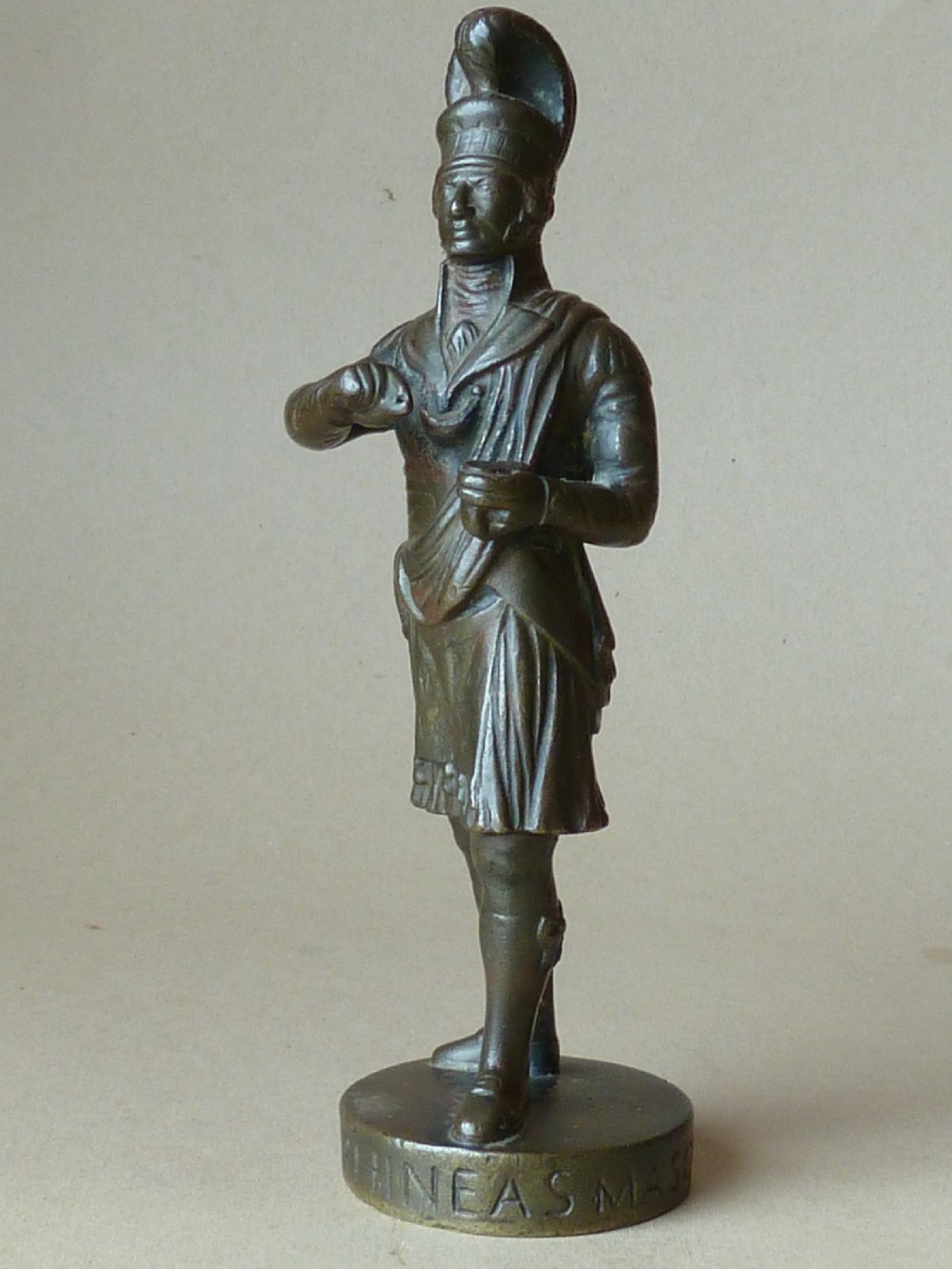 A dark bronze figure depicting a Scottish Highlander in military uniform standing on a circular base inscribed with text. View VII