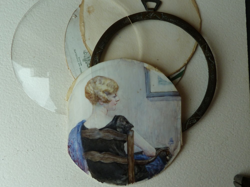 A circular Art Deco hand-painted portrait miniature depicting a 1920s blonde woman in a black dress with a blue wrap, set within an etched circular metal frame. View V