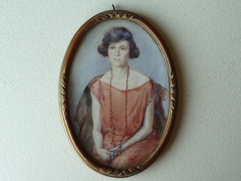 An oval Art Deco portrait miniature depicting a woman with short dark hair in a pinkish-terracotta dress, housed within a twisted metal frame.