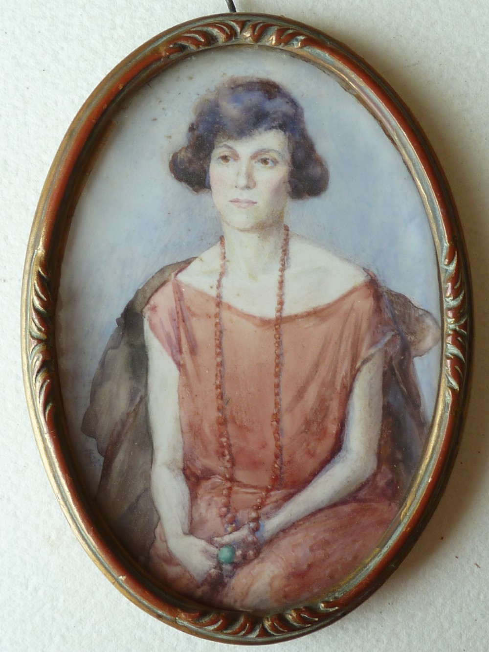 An oval Art Deco portrait miniature depicting a woman with short dark hair in a pinkish-terracotta dress, housed within a twisted metal frame. View II
