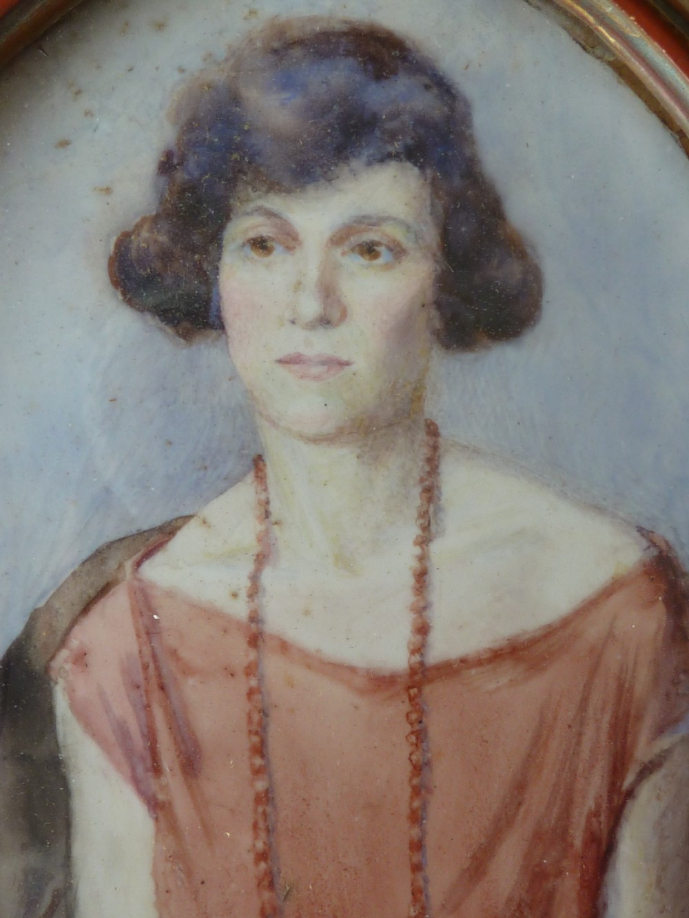 An oval Art Deco portrait miniature depicting a woman with short dark hair in a pinkish-terracotta dress, housed within a twisted metal frame. View III