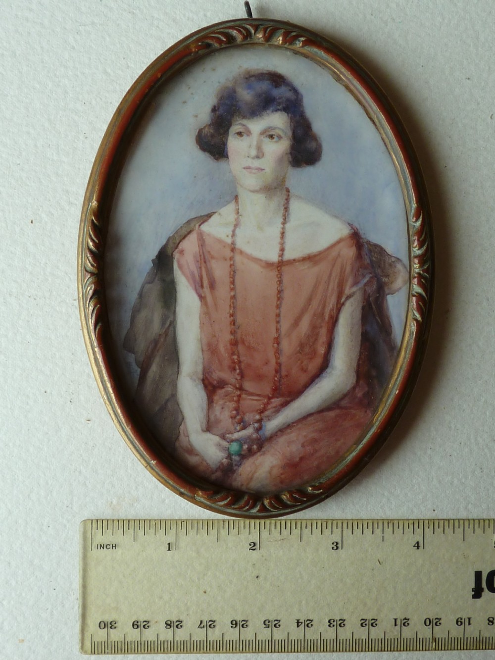 An oval Art Deco portrait miniature depicting a woman with short dark hair in a pinkish-terracotta dress, housed within a twisted metal frame. View VII