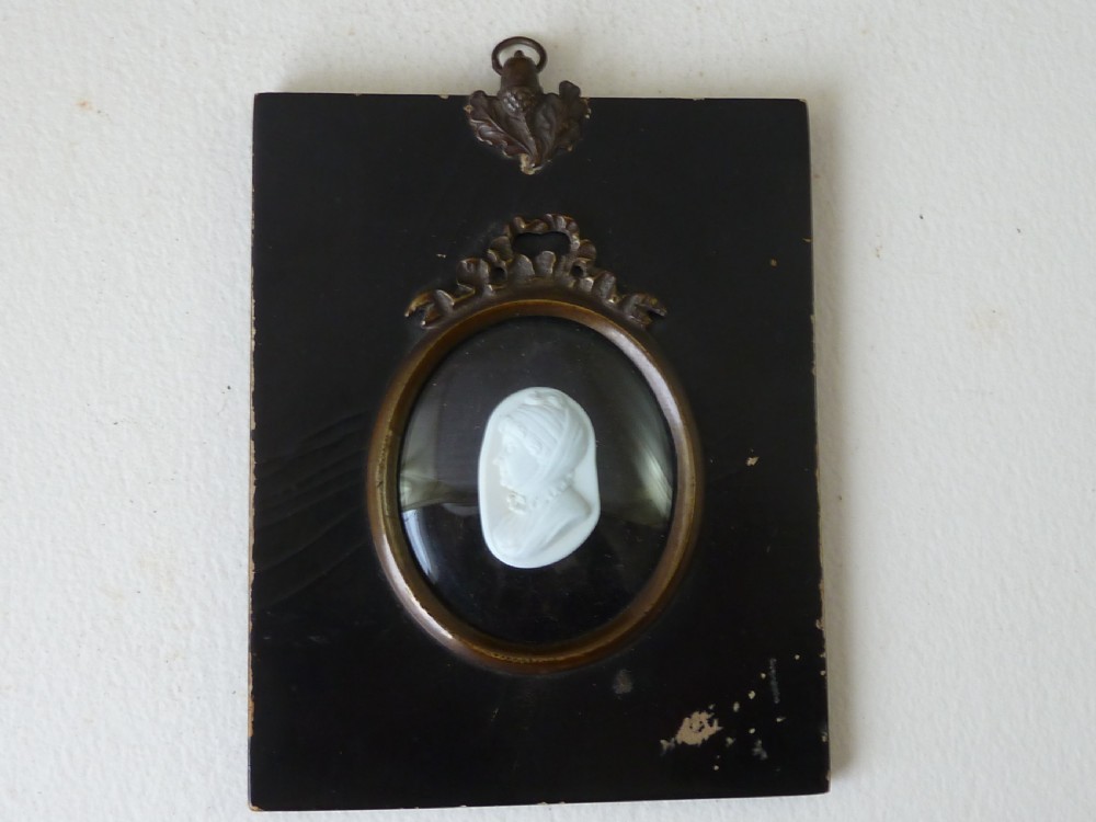 A Georgian oval white glass paste profile bust miniature mounted in a brass bezel on a rectangular black ebonised wooden plaque with a brass acorn hanger.