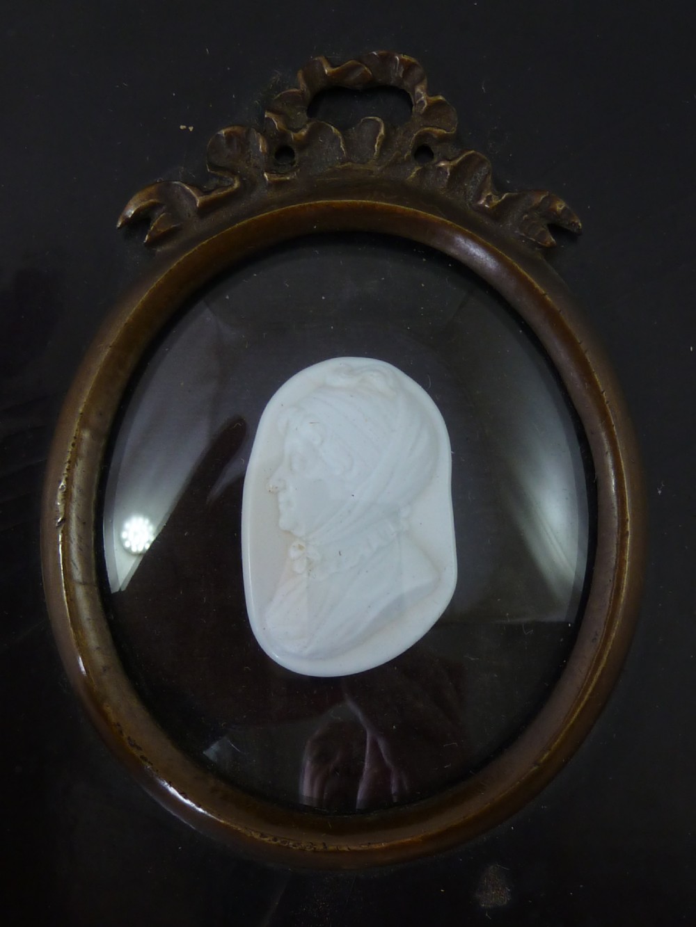A Georgian oval white glass paste profile bust miniature mounted in a brass bezel on a rectangular black ebonised wooden plaque with a brass acorn hanger. View II