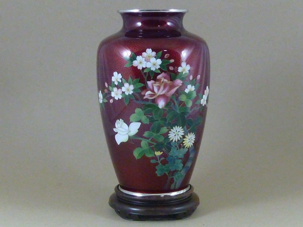 A red Japanese ginbari enamel vase decorated with floral silver wire motifs set upon a dark carved wooden base.
