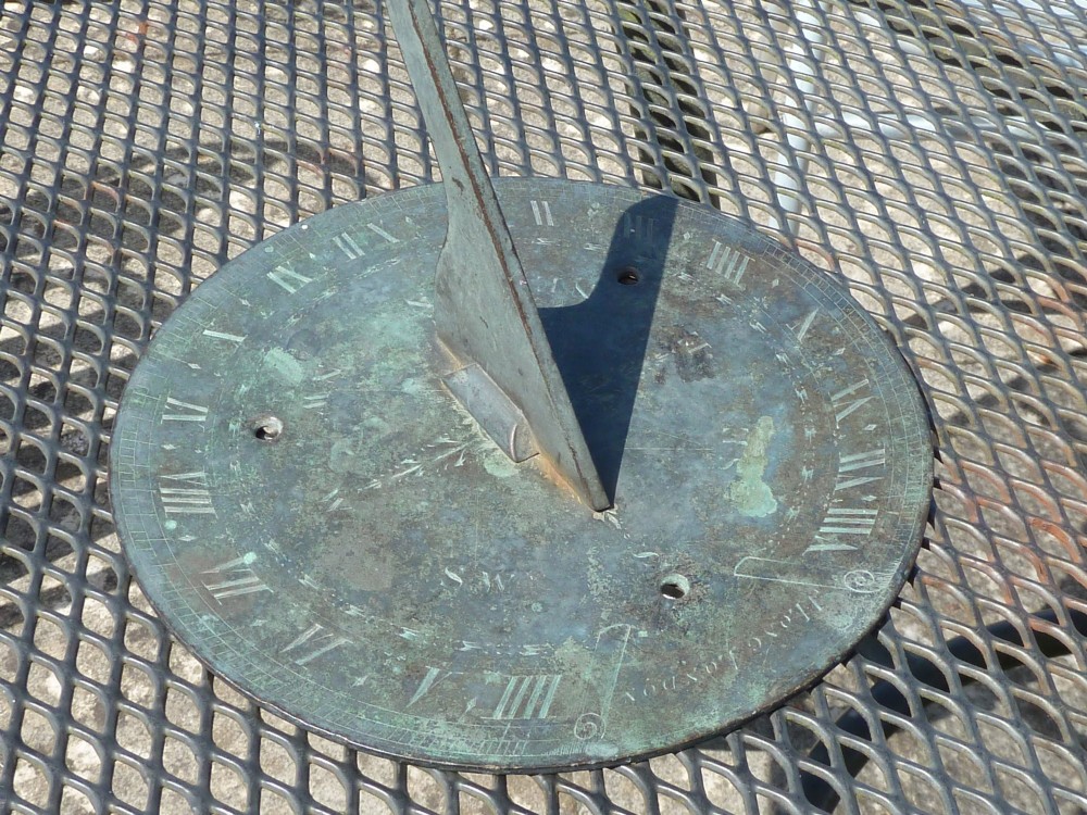 A circular weathered bronze sundial plate with a central triangular gnomon, engraved Roman numerals, and a green verdigris patina. View II