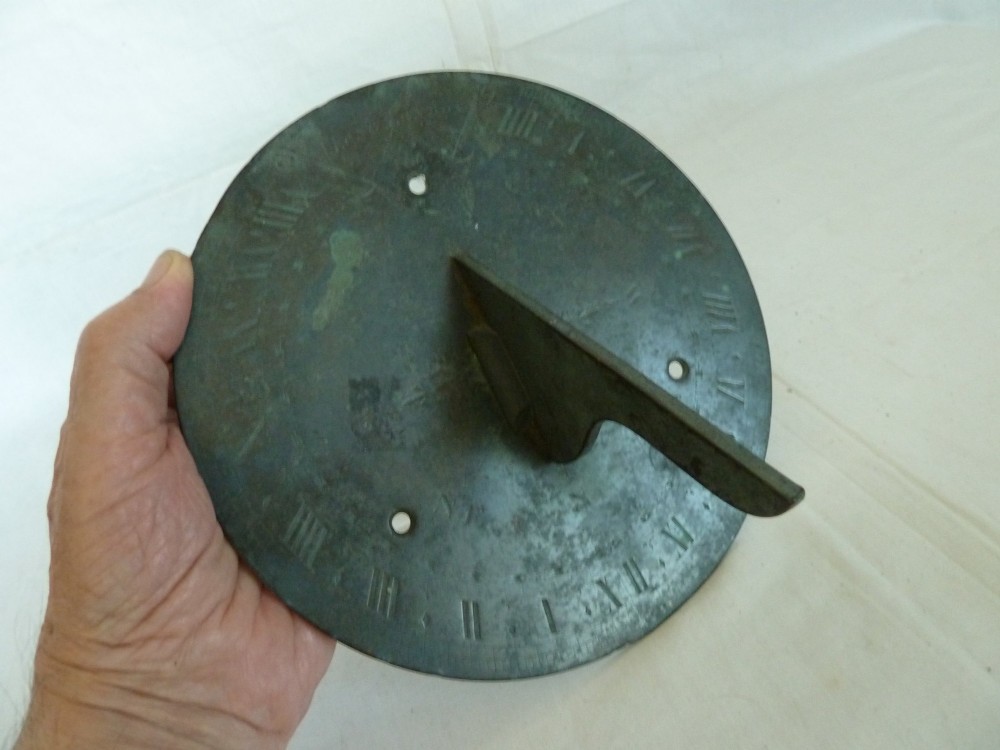 A circular weathered bronze sundial plate with a central triangular gnomon, engraved Roman numerals, and a green verdigris patina. View VI