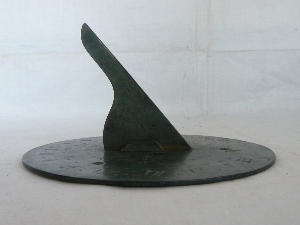 A circular weathered bronze sundial plate with a central triangular gnomon, engraved Roman numerals, and a green verdigris patina. View IX