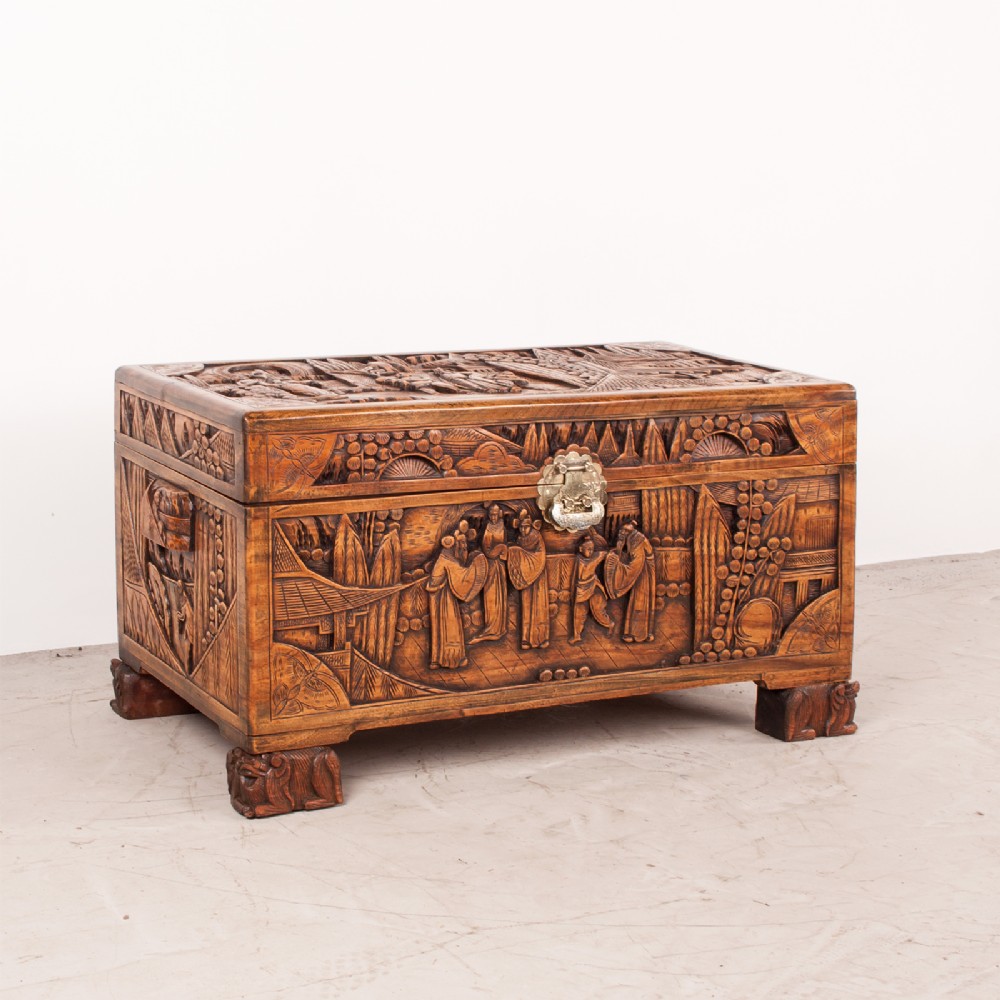 A warm brown carved camphorwood storage chest featuring relief carvings of figures and landscapes with a central brass latch.