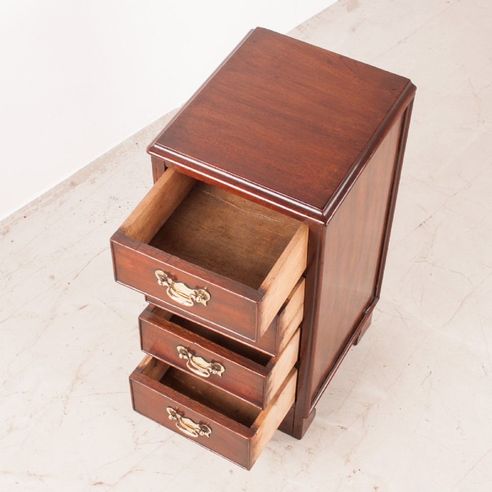 A narrow dark brown mahogany chest featuring four stacked drawers with polished brass drop handles and a shaped bracket foot apron. View V