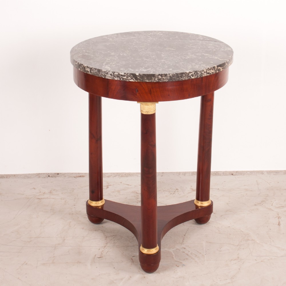 A 19th-century French mahogany gueridon side table with a round grey marble top, three cylindrical legs with gilt mounts, and a tri-form concave base.