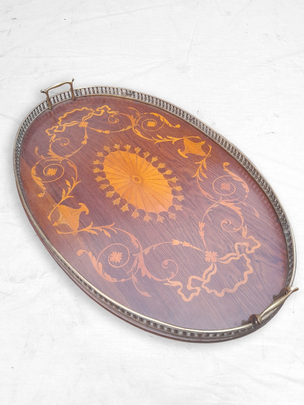 An oval Edwardian mahogany tray featuring intricate satinwood marquetry inlays, a central sunburst medallion, floral scrollwork, and a pierced brass gallery rim with handles.