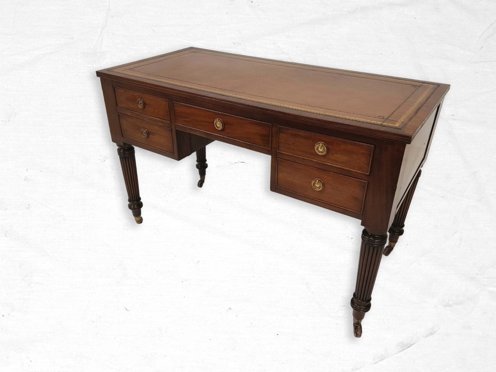 A mahogany writing table featuring five drawers, a brown leather inset surface with gold tooling, reeded turned legs, and brass castors.