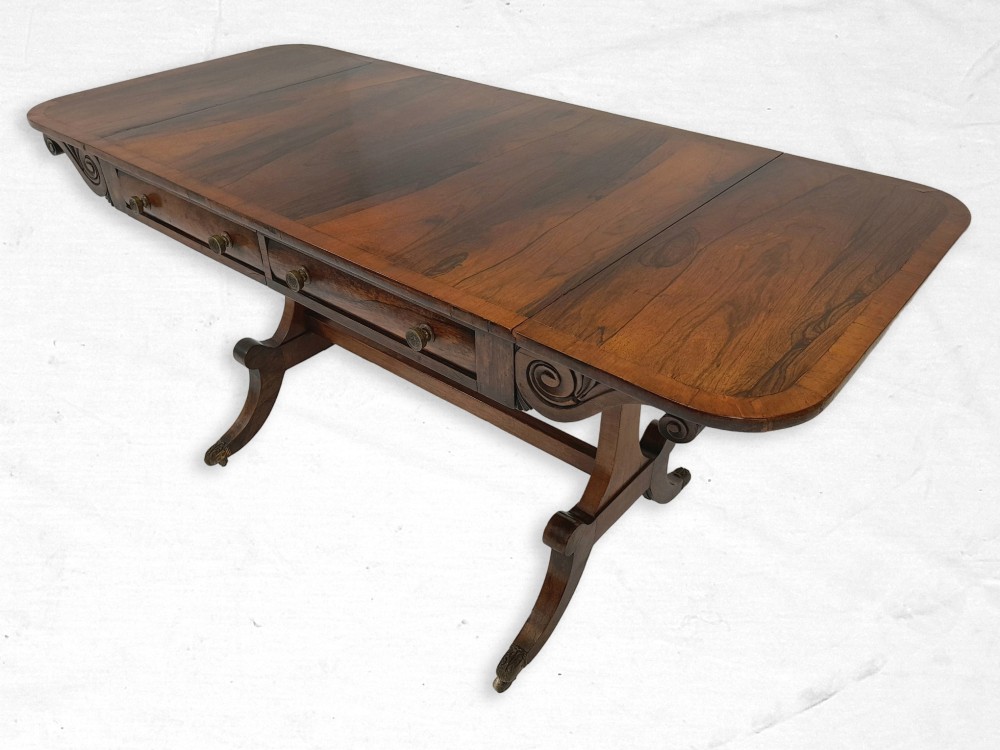A Regency rosewood sofa table featuring drop-leaf flaps, frieze drawers with brass handles, carved scroll supports, and splayed legs on brass castors.